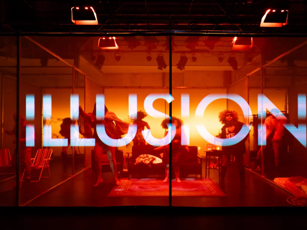 Blick durch ein feines Netz auf eine in Orange-Rot getauchte Bühne, auf der mehrere Personen tanzen. Auf das Netz ist das Wort „ILLUSION" projiziert.