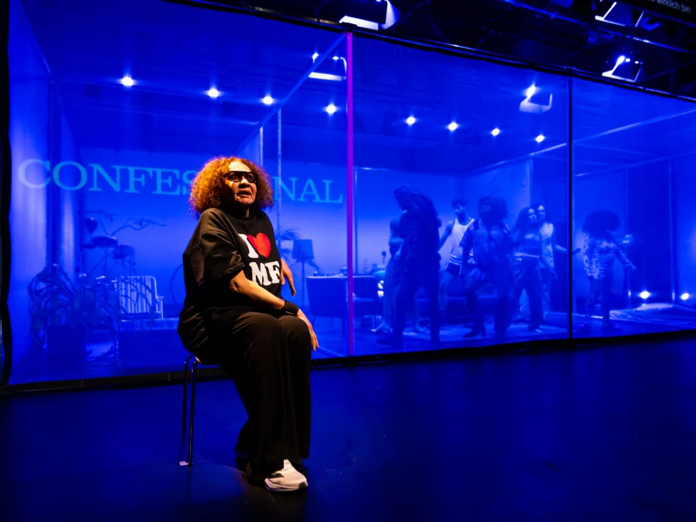 Blick durch eine Glasscheibe auf eine Bühnenszene mit mehreren Personen in einem wohnzimmerartigen Raum. Auf das Glas ist ein englischsprachiger Text projiziert: „Verônika hopes she didn't leave her red wig behind, because her character should be red-haired."