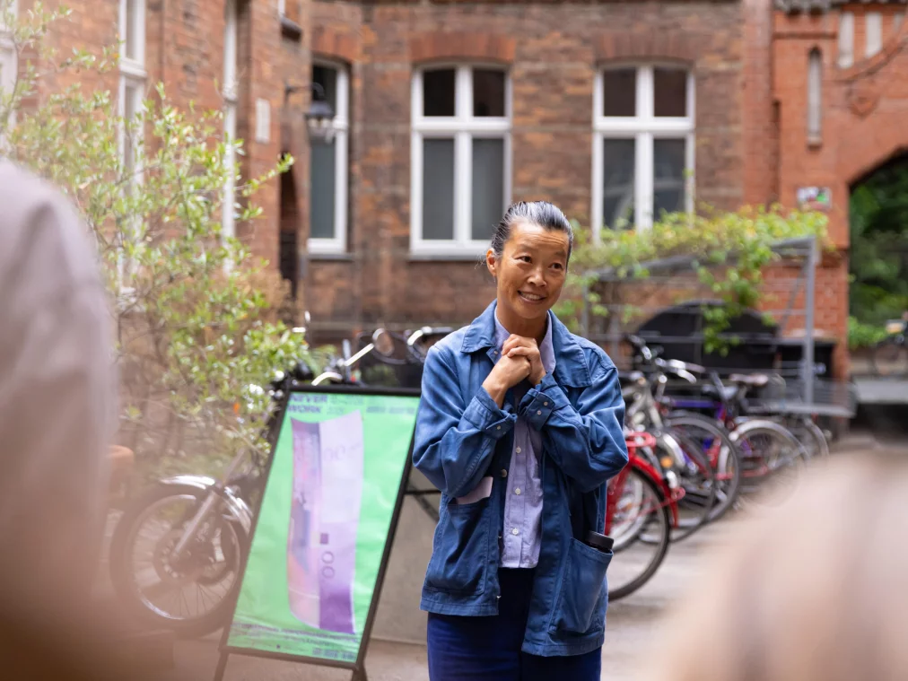 Eine Person in blauer Jeansjacke steht lächelnd vor einem Backsteingebäude mit Fahrradständer und spricht zu einer Gruppe. Links ein Aufsteller mit Veranstaltungsplakat.