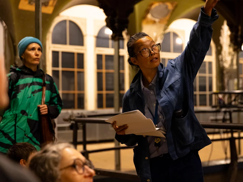 Eine Person mit Brille und blauer Jacke hält Papiere in der Hand und streckt einen Arm aus, während sie spricht. Im Hintergrund stehen weitere Personen in einem historischen Saal mit Bogenfenstern.