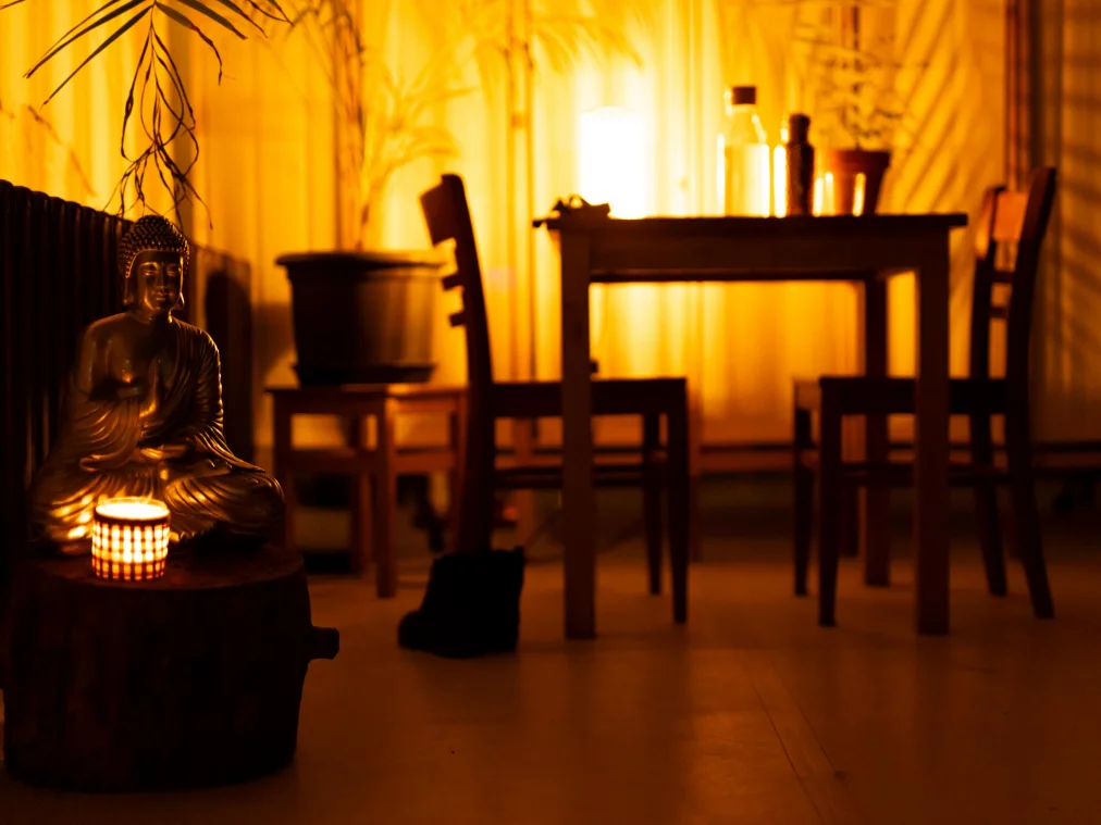 Stillleben in warmem Kerzenlicht: eine Buddha-Figur mit Teelicht auf einem Holzsockel, dahinter Stühle und ein Tisch mit Flaschen, im Hintergrund ein beleuchteter Vorhang.