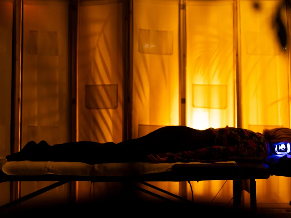 Eine Person liegt als Silhouette auf einer Massageliege; auf ihrem Rücken leuchtet ein kleines blaues Gerät. Im Hintergrund der orangegelb hinterleuchtete Paravent.