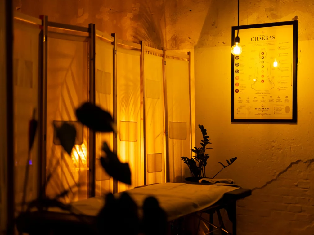 Warmes orangegelbes Licht beleuchtet einen gewölbten Kellerraum mit einem Paravent, einer Massageliege, Topfpflanzen und einer gerahmten Chakra-Grafik an der Wand. Eine Glühbirne hängt von der Decke.