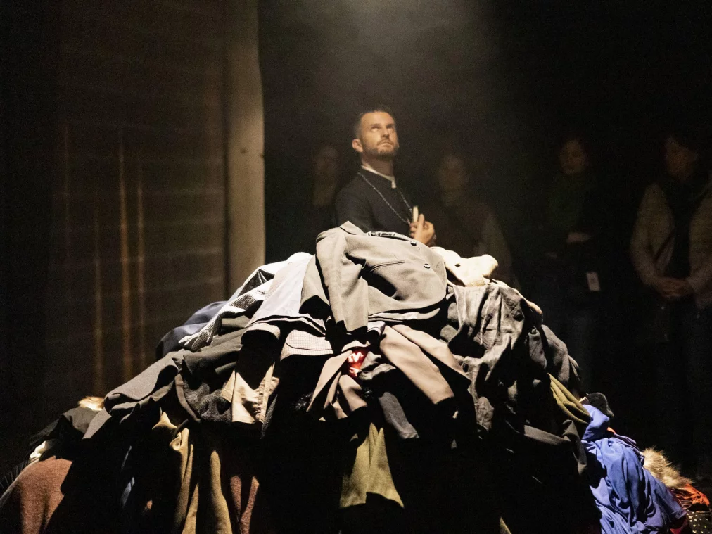 A person stands behind a large pile of heaped clothing, looking upward. Further people observe the scene in the background. Dramatic spotlight.