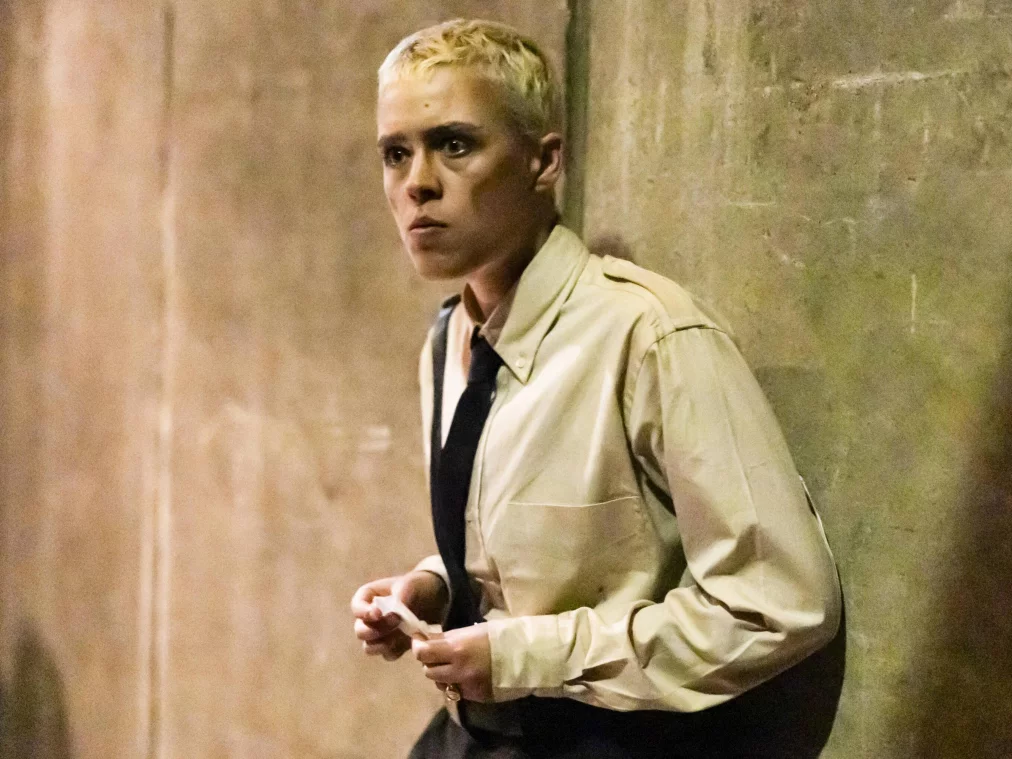 Close-up of a person with short blonde hair and a beige shirt leaning alertly against a weathered concrete wall, glancing to the side.