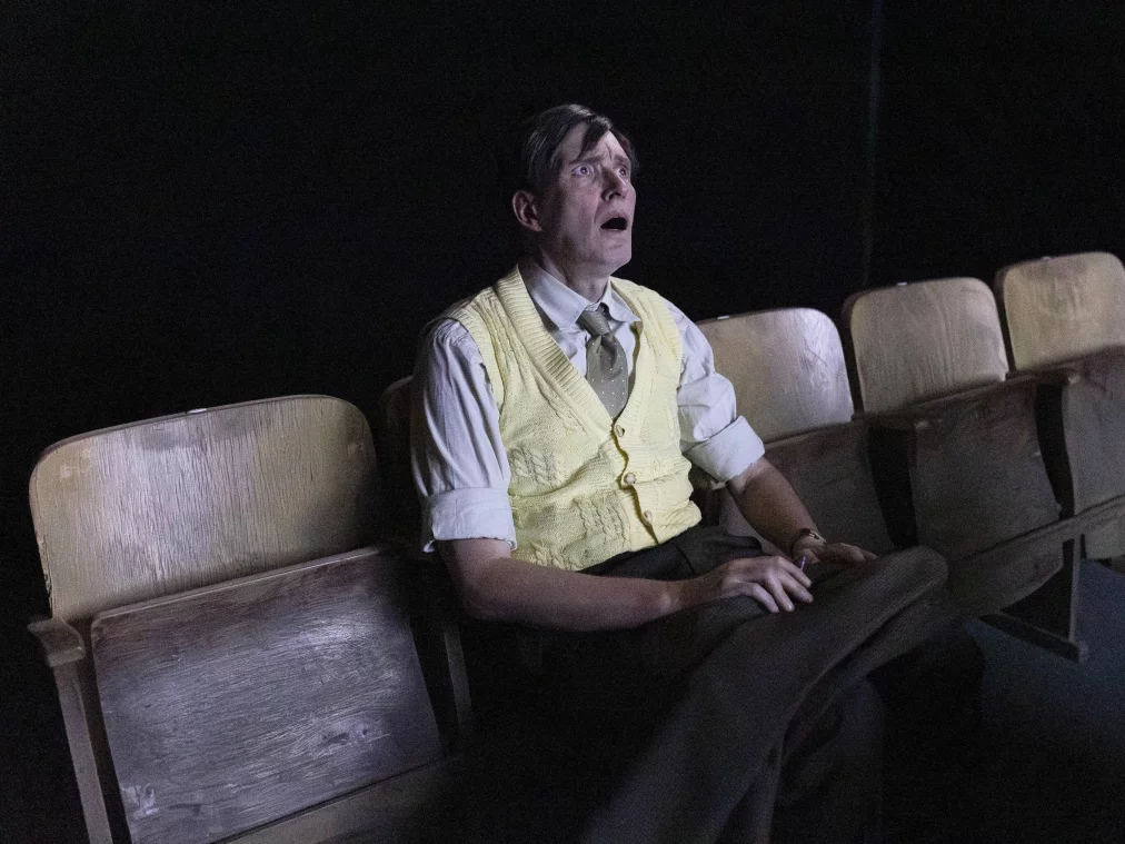 A person in a yellow knit vest and white shirt sits with mouth and eyes wide open on an old wooden folding chair in a row of empty chairs. Dark background.