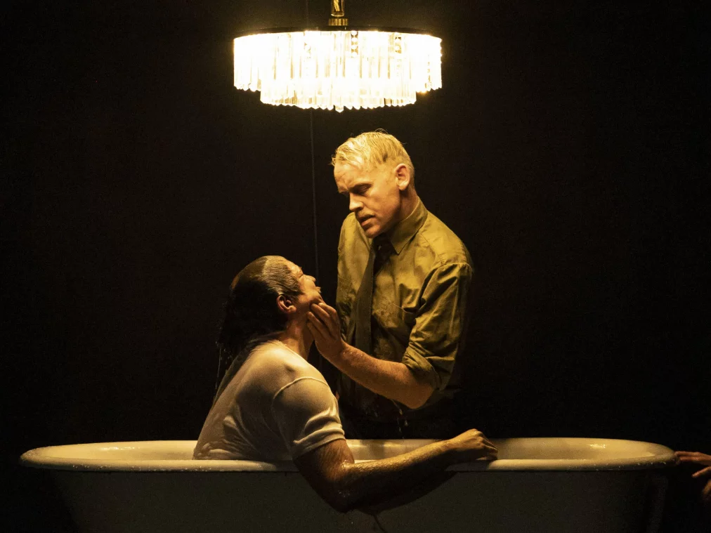 Two people in a dramatic scene at an old-fashioned bathtub beneath a chandelier: one person sits wet in the tub, another leans over them. A third person in uniform is visible at the right edge. Dark background.