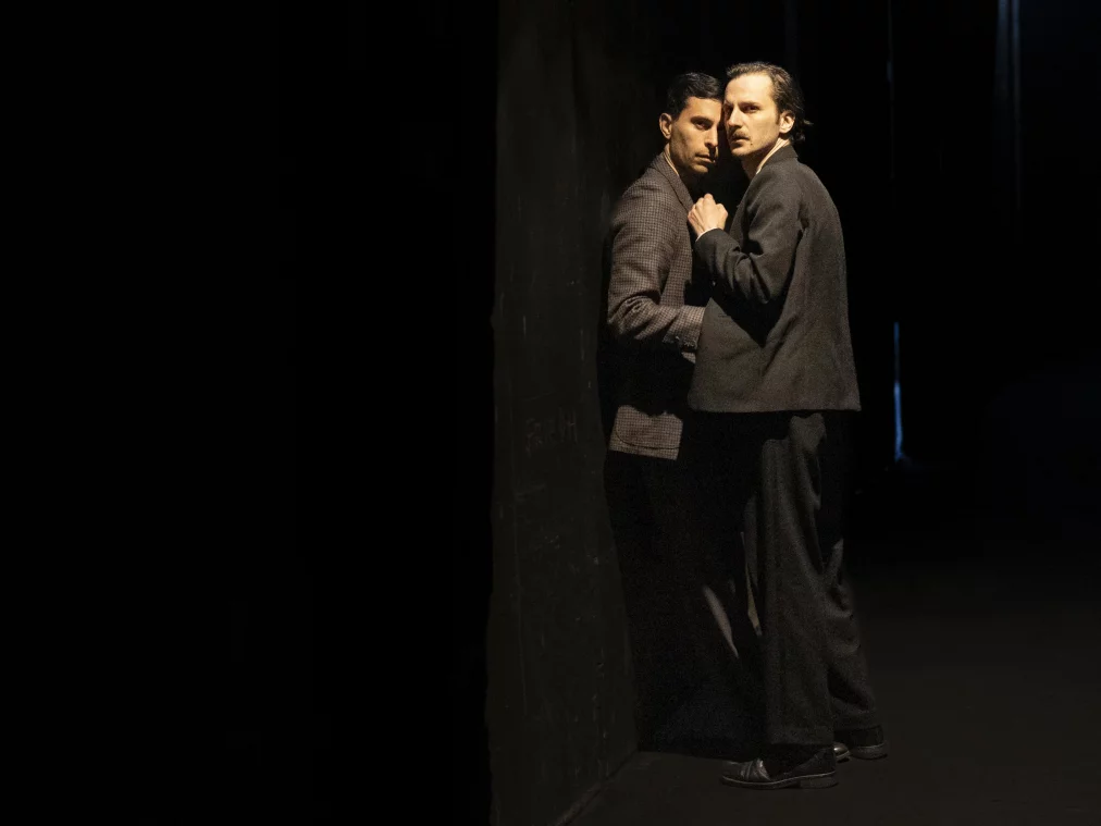Two people in dark suits stand pressed close together against a wall on a dark stage, looking into the camera.