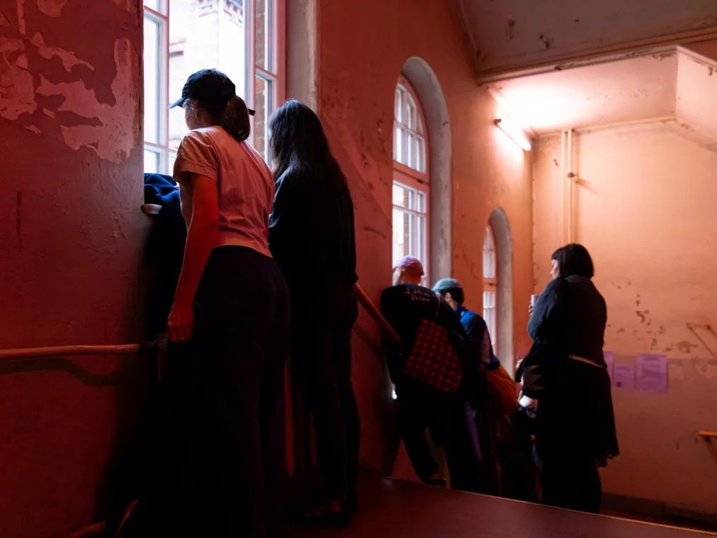 LaurieYoung-AfterWorkTourNo1©MayraWallraff-1Several people stand in a red-lit stairwell with weathered walls, looking out through arched windows.7