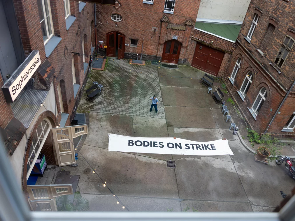 Bird's-eye view of a courtyard at Sophiensæle. A large white banner reading "BODIES ON STRIKE" lies on the ground; a person stands beside it. Brick buildings surround the courtyard.