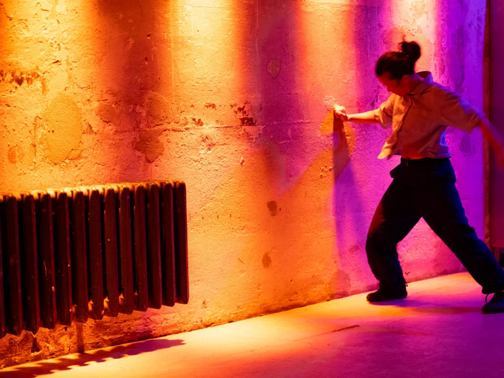 A person in a loose shirt and dark trousers leans dynamically against a wall of weathered masonry. Warm orange-pink stage lighting, an old radiator to the left.