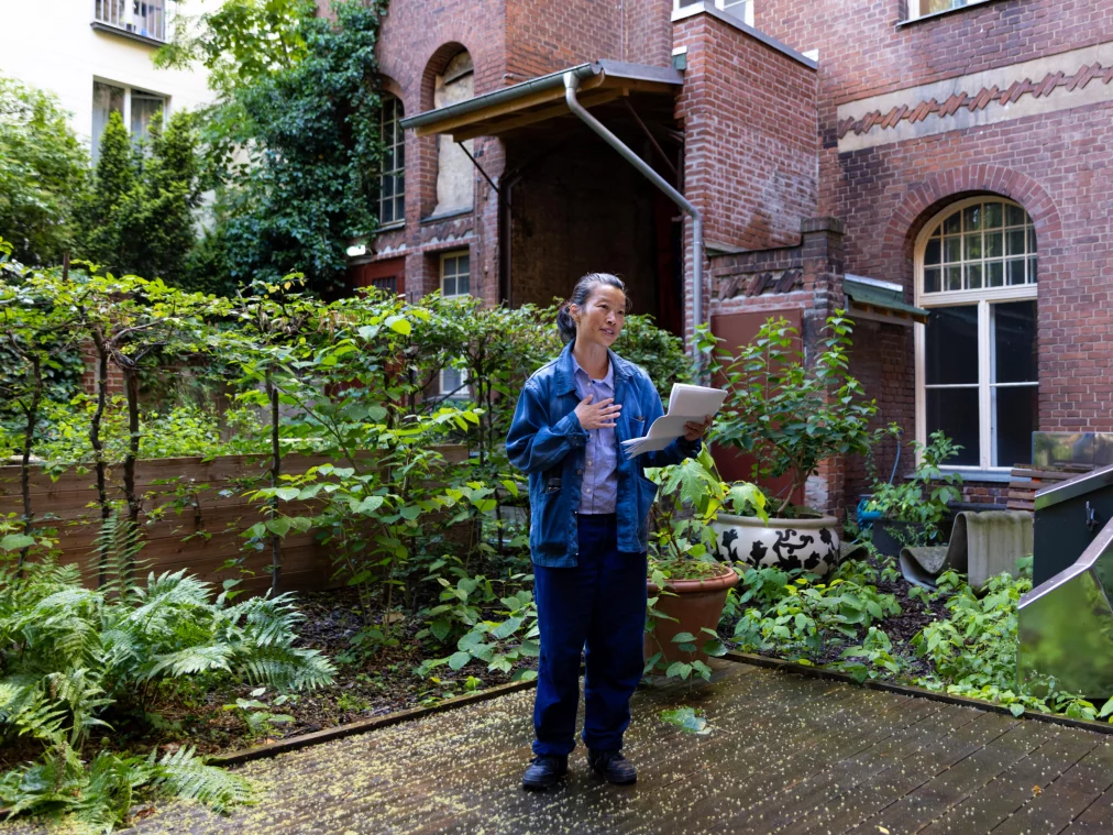 A person in a blue jacket stands in a green courtyard holding papers and speaking. A brick building with arched windows in the background.