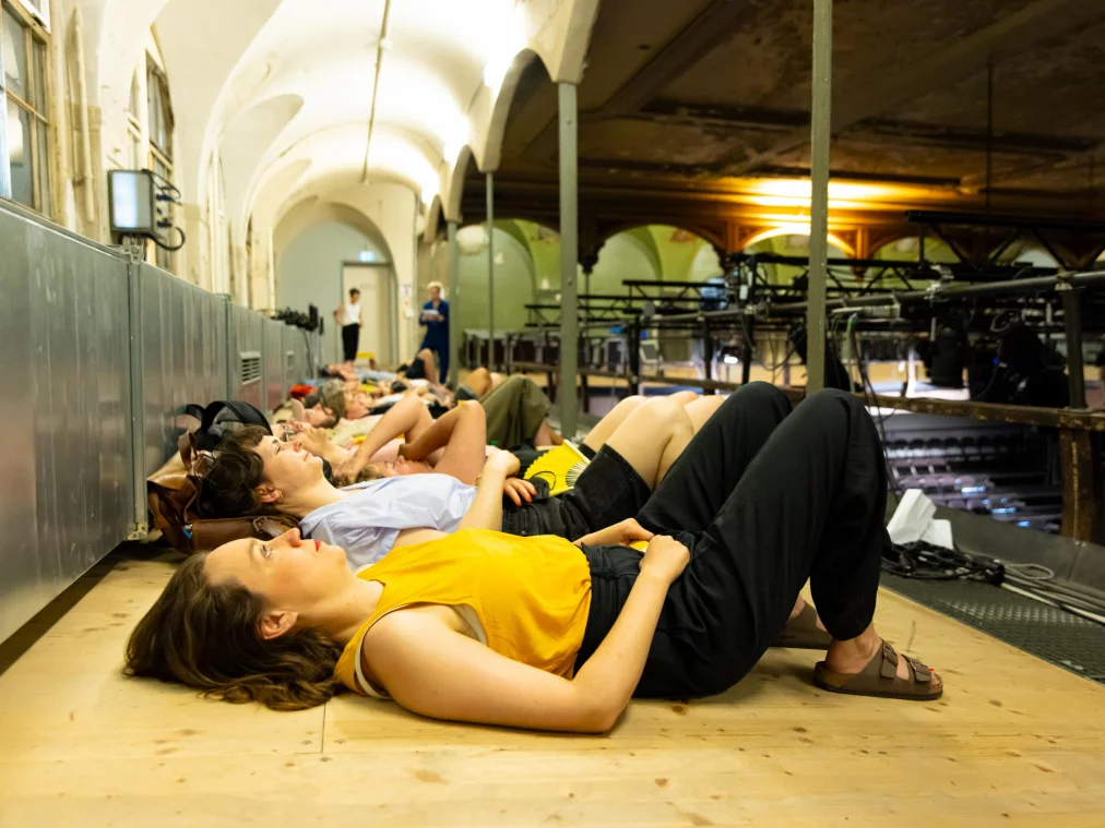 Several people lie side by side on the floor of a historic vaulted hall, looking upward. Two more people are visible standing in the background.