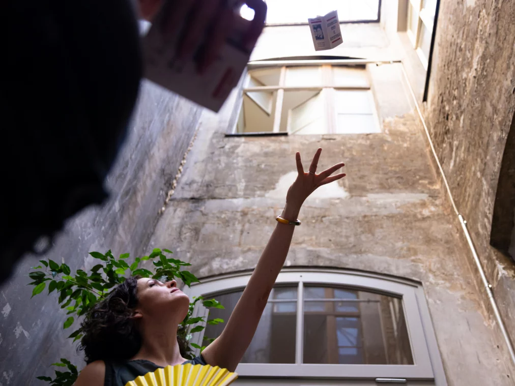 A person throws a booklet into the air in a courtyard; weathered masonry with arched windows in the background. Low-angle perspective.
