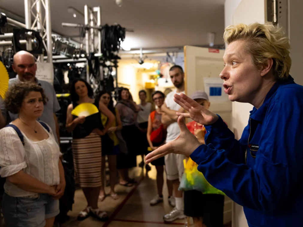 A person in a blue boiler suit speaks with gestures to a group of people in a backstage area with stage technology in the background.