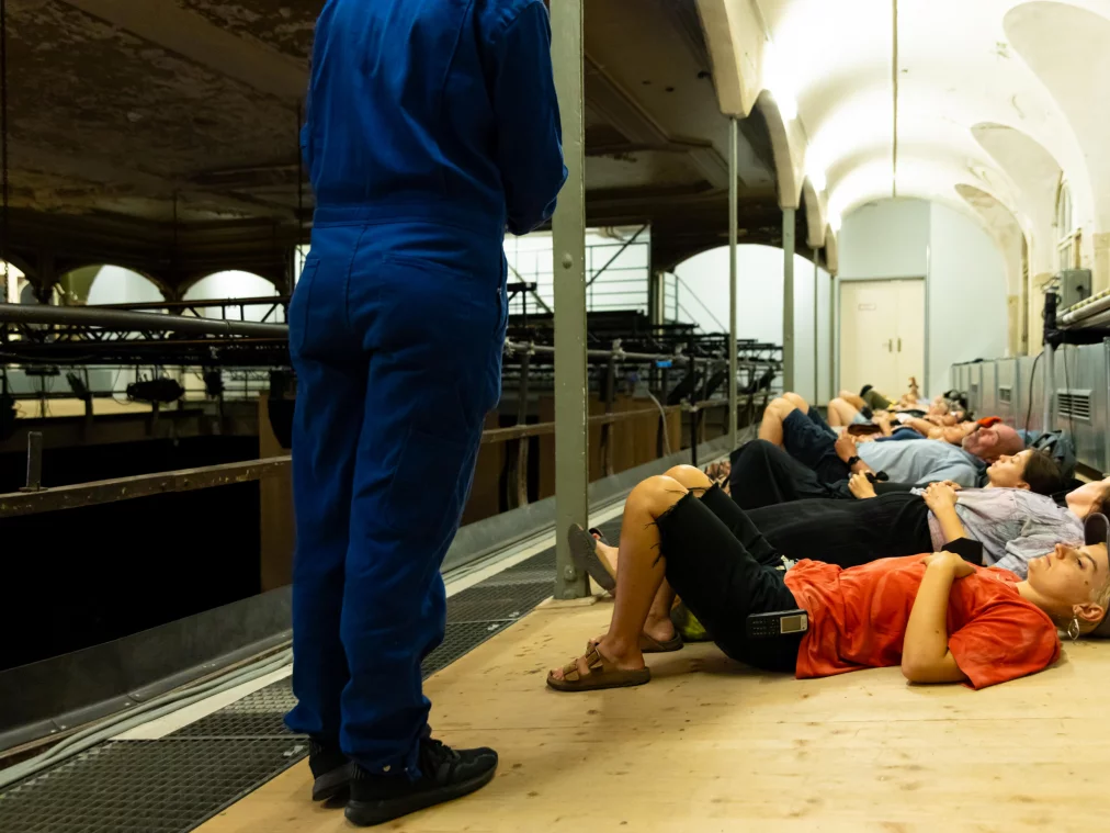 Several people lie side by side on the floor of a historic vaulted hall; a person in a blue boiler suit stands to the left. Stage technology in the background.