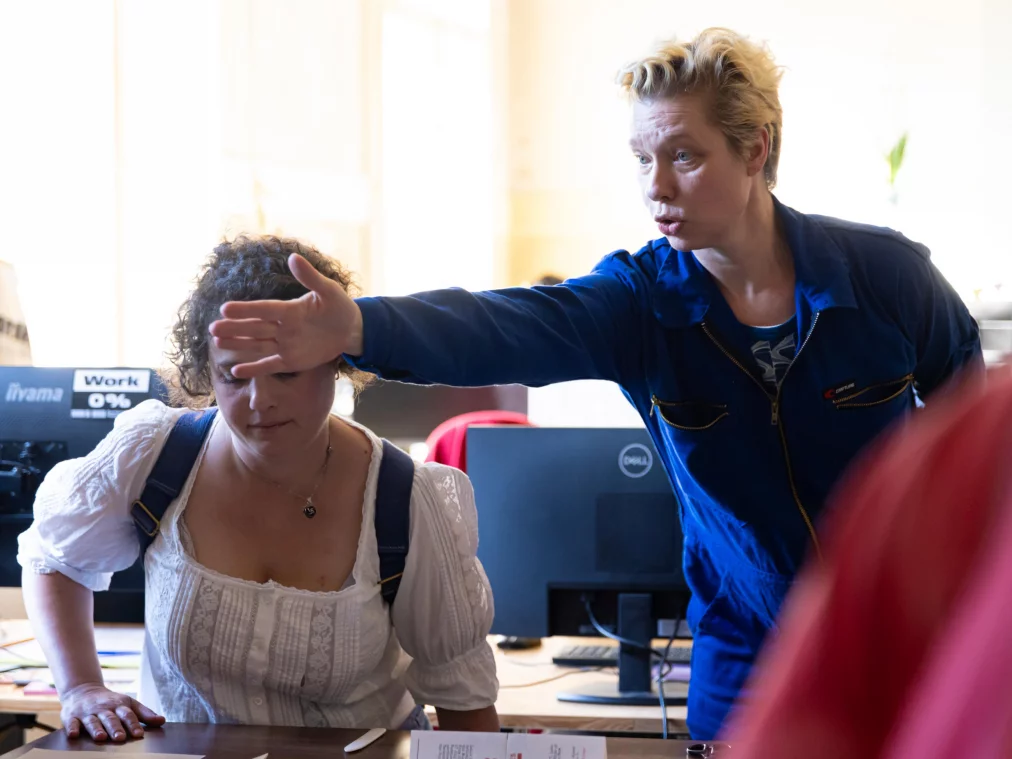Two people in an office: one in a blue boiler suit leans over a seated person and points with an outstretched arm. Computer screens in the background.