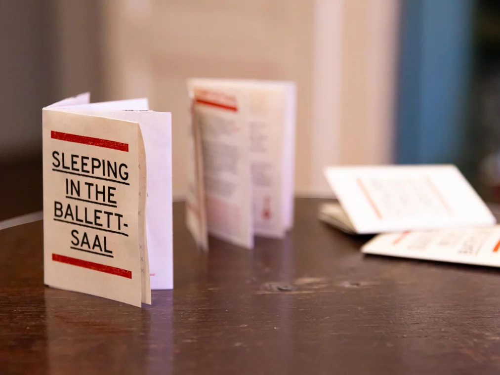Several folded booklets stand and lie on a table; the one in the foreground reads "Sleeping in the Ballettsaal". Blurred background.