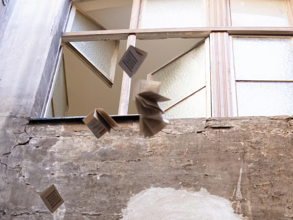 Several folded booklets fly through the air in front of a weathered wall with an old multi-pane window.