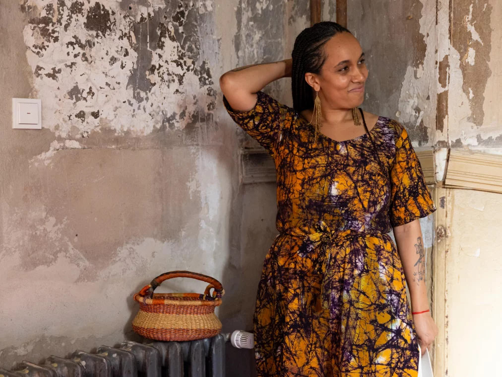 A person in a patterned orange dress and long earrings leans smiling against a weathered wall; beside them a basket on an old radiator.