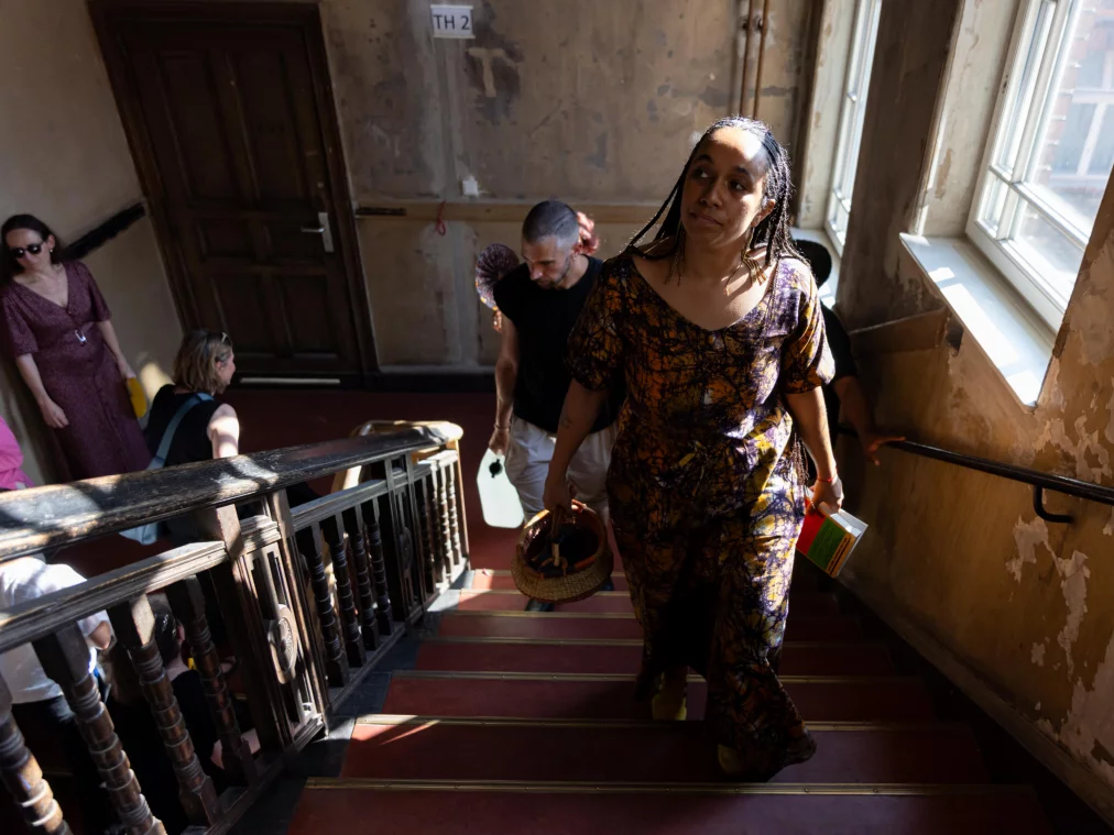 ColleenNdemehFitzgerald-AfterWoA person in a patterned orange dress ascends a historic wooden staircase, holding a basket. Further people follow in the background.rkTourNo3©MayraWallraff-24