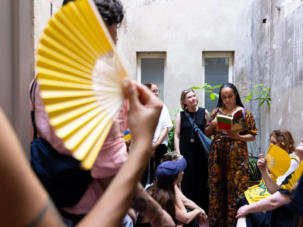 A person in a patterned orange dress reads aloud from a book; a group of people surrounds them, several holding yellow fans. Weathered inner courtyard.