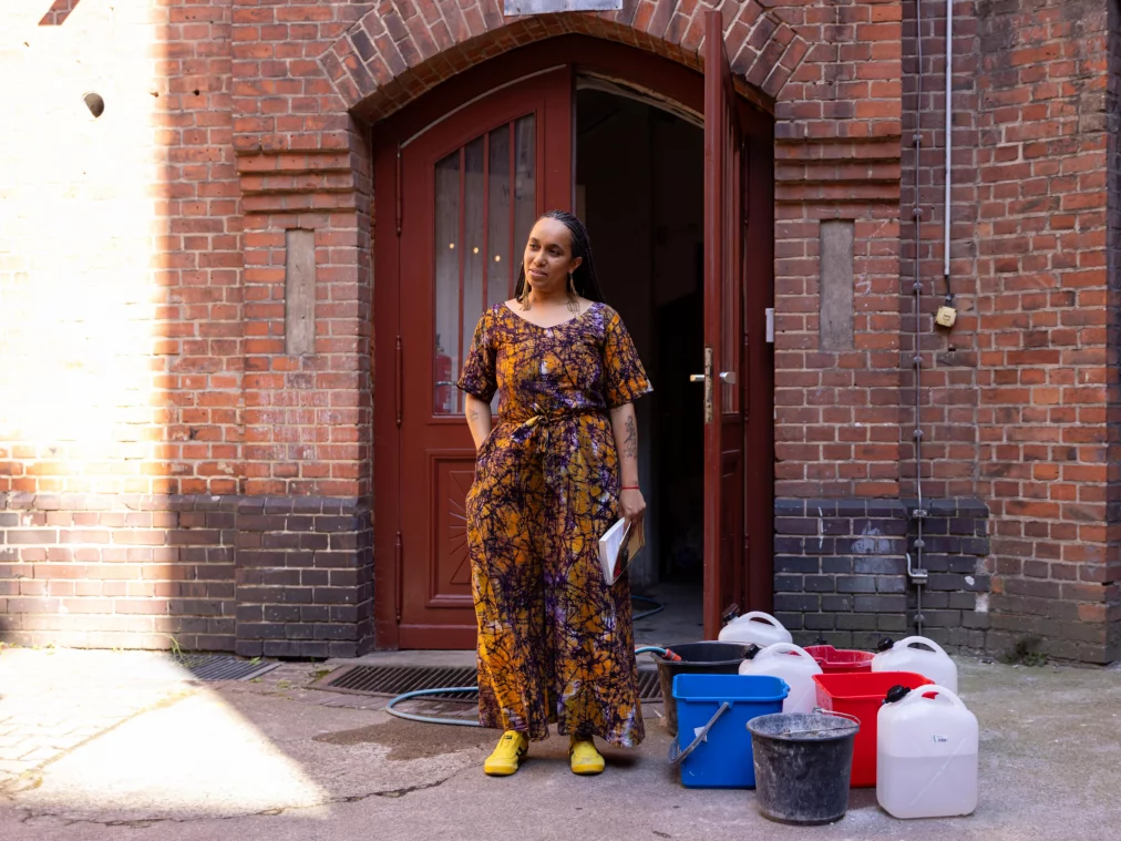 A person in a patterned orange dress and yellow shoes stands in front of a red brick wall with an arched gate. Various buckets and jerrycans are arranged in front of them.