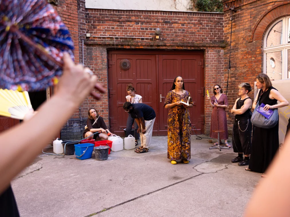 A person in a patterned orange dress reads aloud from a book in a courtyard; several people stand and sit around them. Colourful buckets and jerrycans in the foreground, a red brick wall in the background.