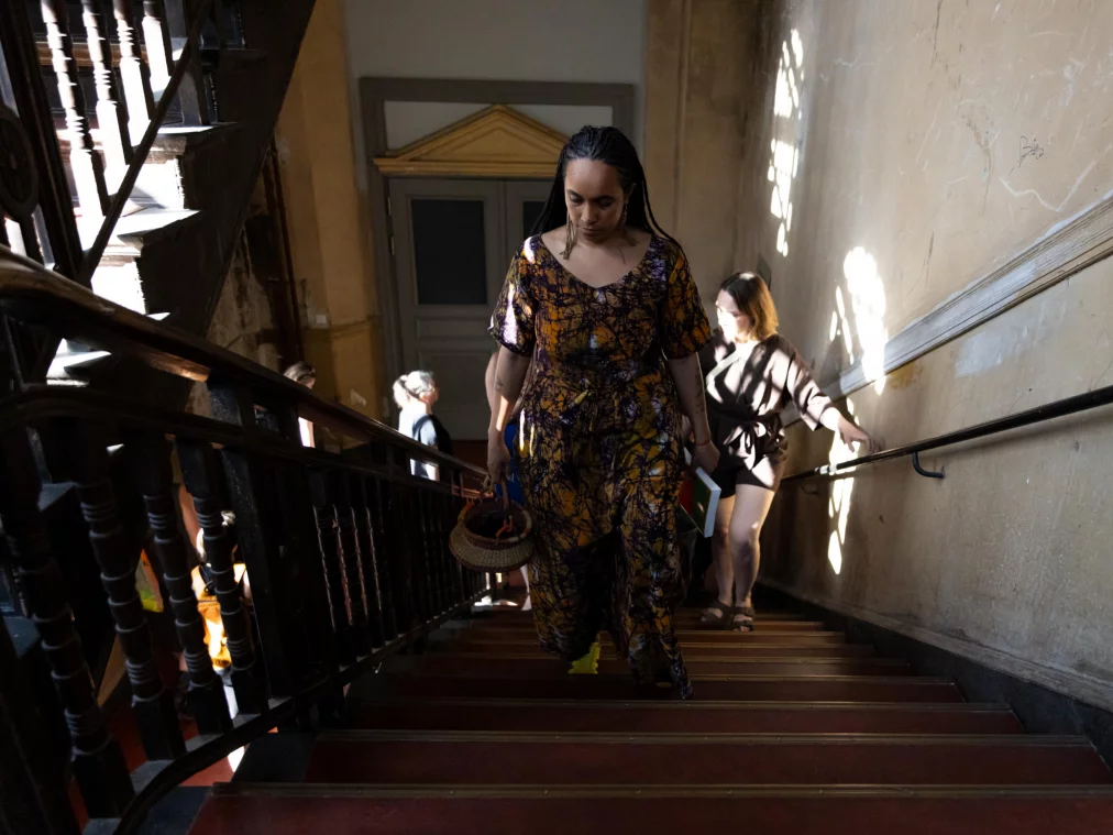 A person in a patterned orange dress ascends a historic wooden staircase, holding a basket and a book. Further people follow behind in sunlight.
