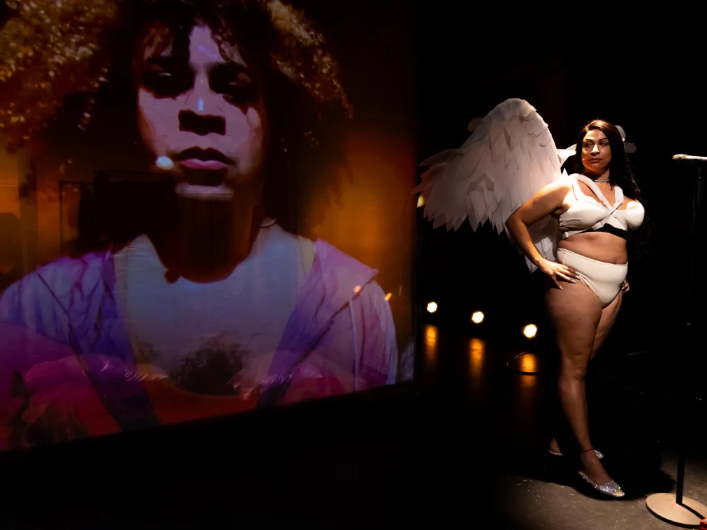 Eine Person in weißem Bustier und Höschen mit großen weißen Flügeln steht auf einer dunklen Bühne neben einem Mikrofonstativ. Links davon eine großformatige Videoprojektion eines Gesichts.
