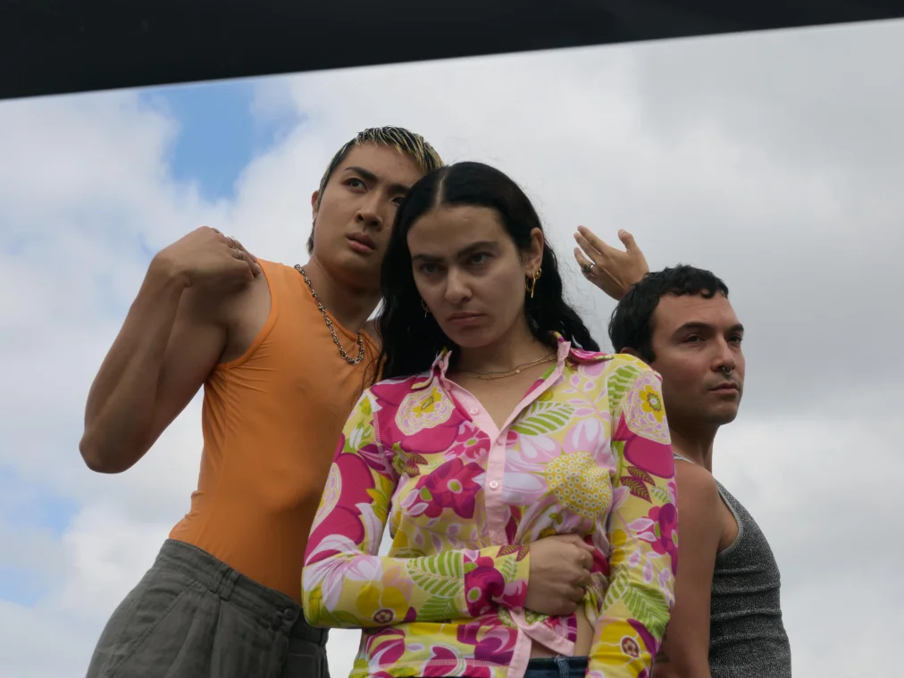 Three people stand close together against a cloudy sky, photographed from below. In the middle, a person in a colourful floral shirt looks down seriously. On the left, a person in an orange top leans their head against them and raises a fist to their shoulder. On the right stands a person in a grey top, with a hand raised behind them.