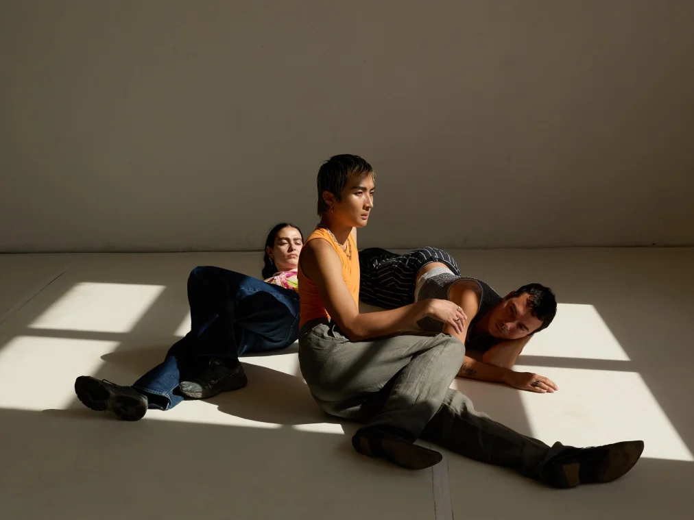 Three people on the floor of a bright room, with slanting sunlight falling through a window. In front, a person in an orange top and grey trousers sits looking to the side, one hand resting on the hip of a person lying on their side behind them. On the left, a person in jeans leans against the seated person's back with eyes closed.