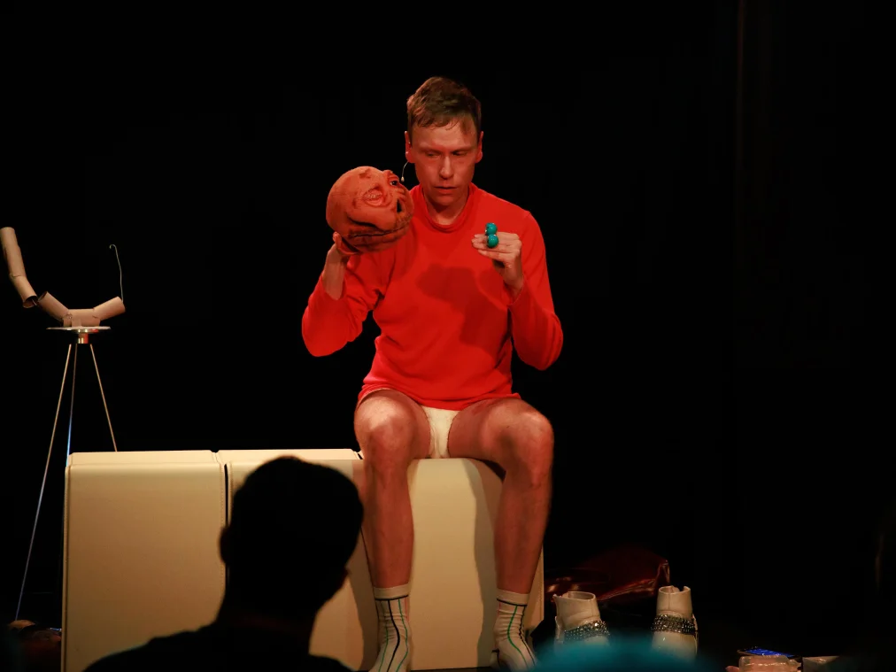 Eine Person in rotem Pullover, weißer Unterhose und weißen Socken sitzt auf einem weißen Quader. In einer Hand hält sie eine orangefarbene Handpuppe, in der anderen zwei türkisfarbene Kugeln, und blickt dabei auf die Puppe. Neben ihr stehen weiße Stiefel mit Ketten, links eine Skulptur aus Papprollen auf einem Stativ.