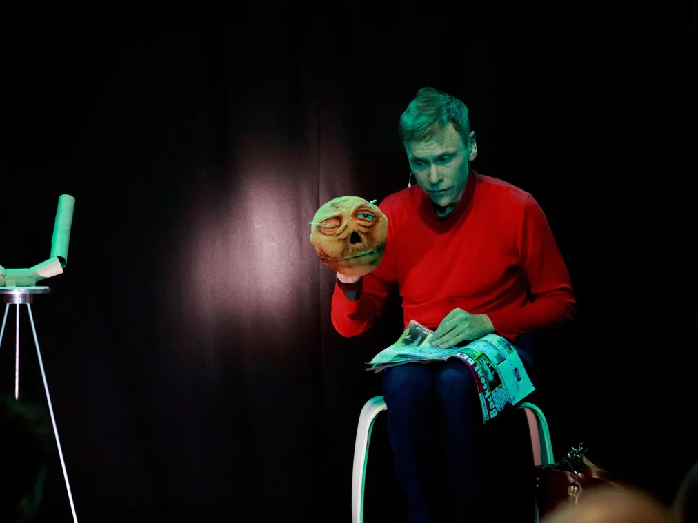 Eine Person in rotem Pullover sitzt auf einem weißen Hocker und blättert in einer Zeitschrift auf ihrem Schoß. Mit der anderen Hand hält sie eine Handpuppe hoch: einen runzligen, orangefarbenen Kopf mit einem halb geschlossenen Auge. Links steht eine Skulptur aus Papprollen auf einem Metallstativ, das Licht ist grünlich.