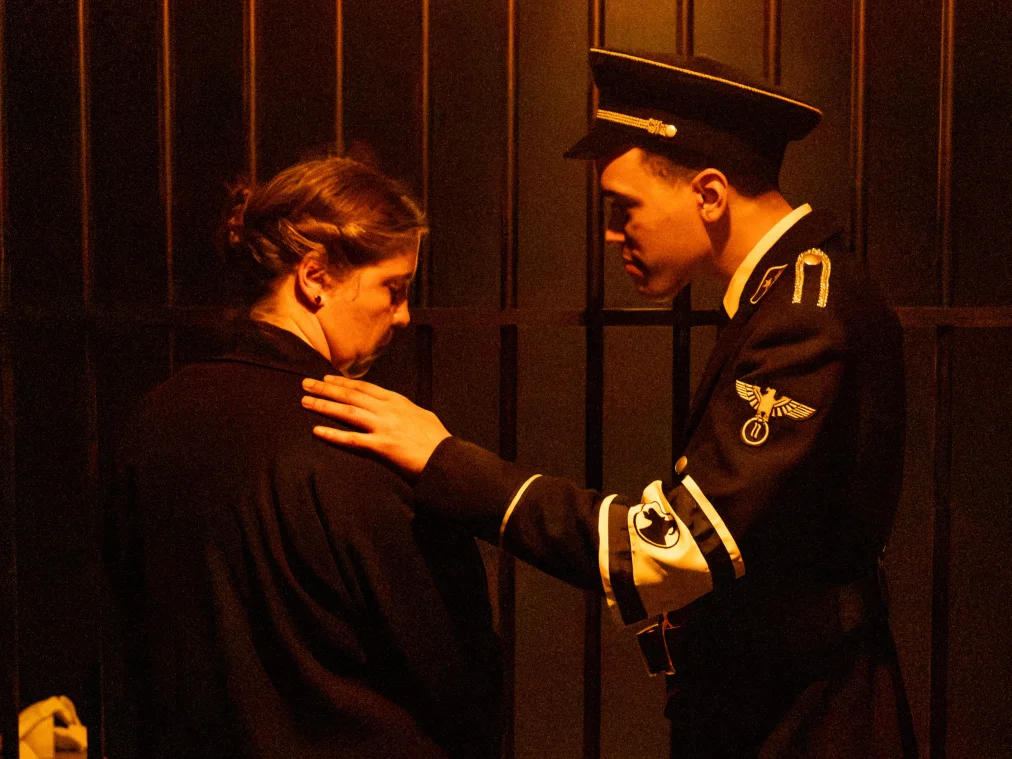 or dunklen Gitterstäben legt eine Person in dunkler Uniform im Stil der NS-Zeit mit Schirmmütze und weißer Armbinde einer Person im schwarzen Mantel die Hand auf die Schulter. Beide senken den Blick. Gedämpftes, warmes Licht.