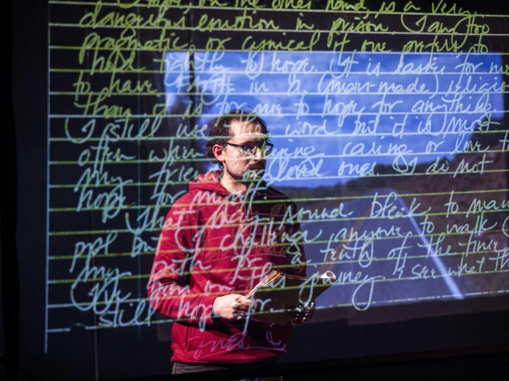 Eine Person mit Brille und rotem Kapuzenpullover steht vor einer großen Projektion und hält einen aufgeschlagenen Aktendeckel. Auf die Fläche und auf die Person projiziert: englische Handschrift in hellgrüner Schreibschrift auf linierten Zeilen, dahinter ein blau schimmerndes Landschaftsbild.