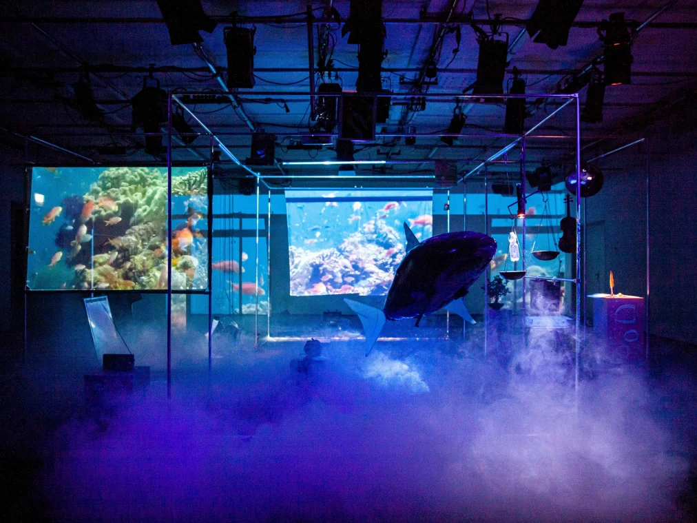 Bühnenbild in blauem Licht und dichtem Bodennebel: In einem Gerüstrahmen hängt ein großer aufblasbarer Hai. Links und in der Mitte zeigen zwei Projektionsflächen Korallenriffe mit Fischen. Rechts hängen eine Balkenwaage und eine Geige, daneben ein Podest mit einer Feder. Über allem die offene Technikdecke mit Scheinwerfern.