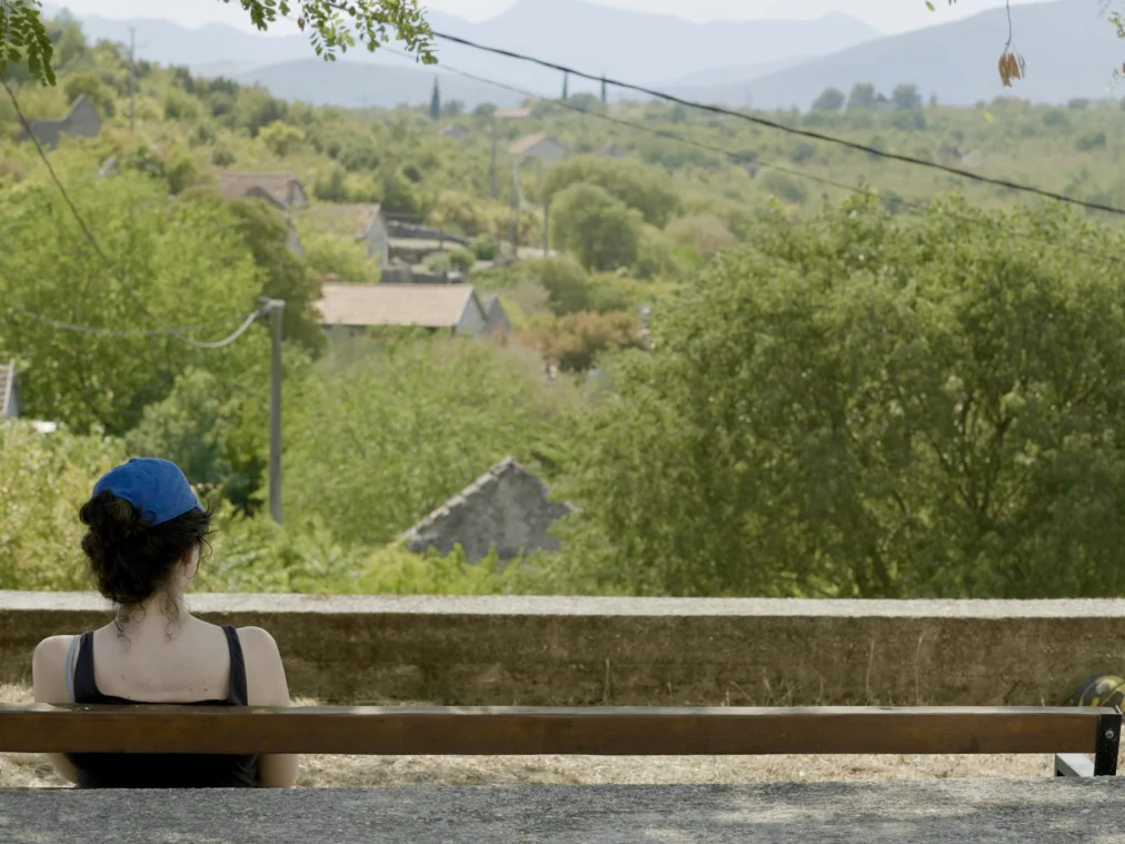A person in a blue cap and dark top sits alone, seen from behind, on a long wooden bench in front of a low wall. The view stretches across green hills with scattered houses, utility poles and power lines to hazy mountains. Two balls and a stone lie beside the wall.