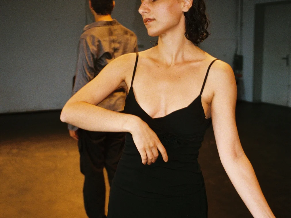 Eine Person in schwarzem Trägerkleid steht im Vordergrund, einen Arm waagerecht vor der Brust angewinkelt, den anderen schräg nach unten gestreckt, der Blick zur Seite. Im Hintergrund steht eine Person in grauem Hemd mit dem Rücken zur Kamera. Ein Scheinwerfer wirft warmes Licht in den leeren Raum.