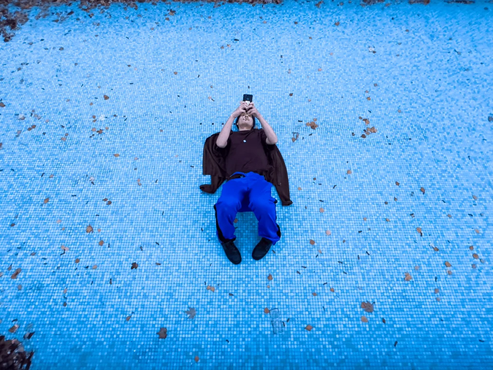 Eine Person liegt auf dem Rücken in einem leeren Schwimmbecken mit hellblauen Mosaikfliesen, auf denen vereinzelt Herbstlaub liegt. Die Person hält ein Smartphone mit beiden Händen über das Gesicht und hat eine Zigarette im Mund. Sie trägt ein dunkelbraunes T-Shirt, eine kräftig blaue Trainingshose mit schwarzem Seitenstreifen und schwarze Stiefel. Unter ihr ist eine braune Jacke ausgebreitet.