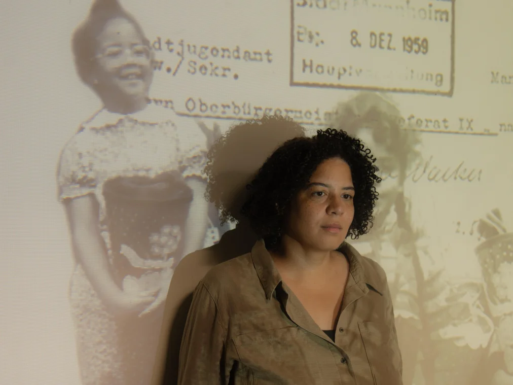 A person leans against a wall, gazing thoughtfully past the camera, arms hanging loosely. They wear a beige-brown shirt with rolled-up sleeves and black trousers. Historical black-and-white photos of two children holding school cones and an official letter stamped “Stadt Mannheim, 8. DEZ. 1959” are projected onto the wall and their body.