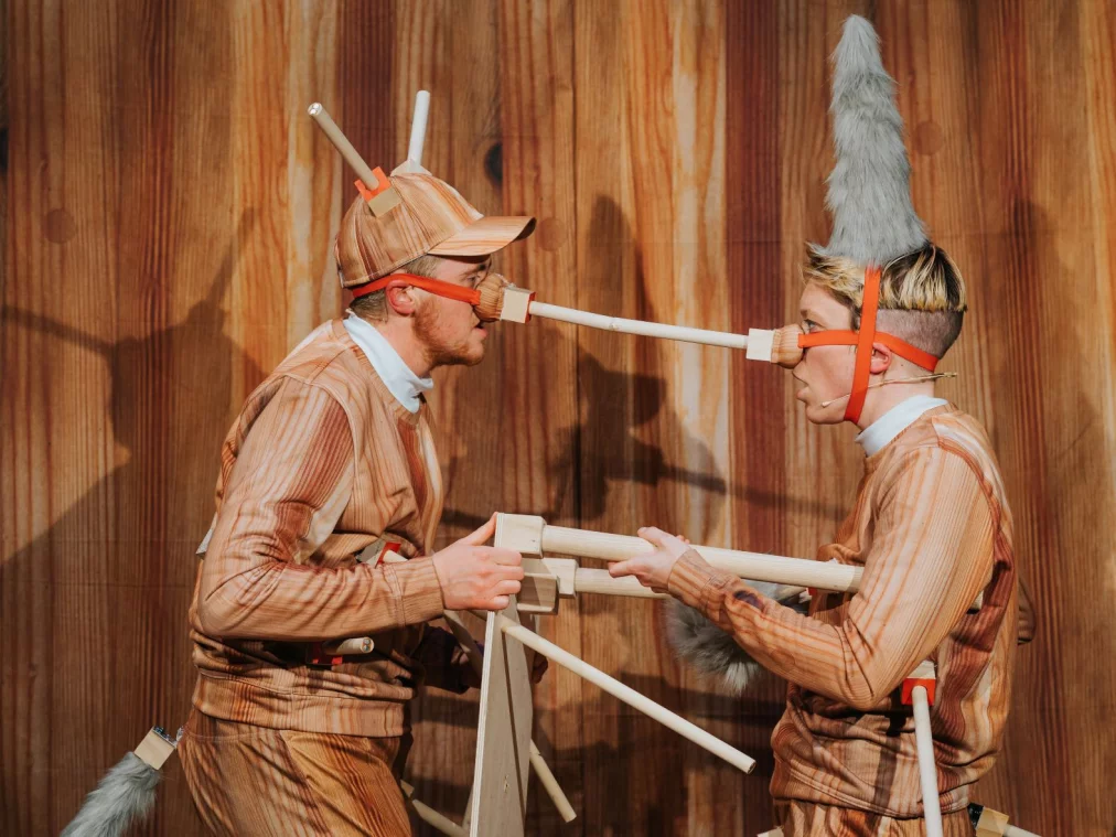Two performers: Ivor MacAskill (he/him) and Rosana Cade (they/them), are shown in profile from the waist up, facing each other. They wear fake wooden tracksuits and caps with real wooden appendages protruding from them.