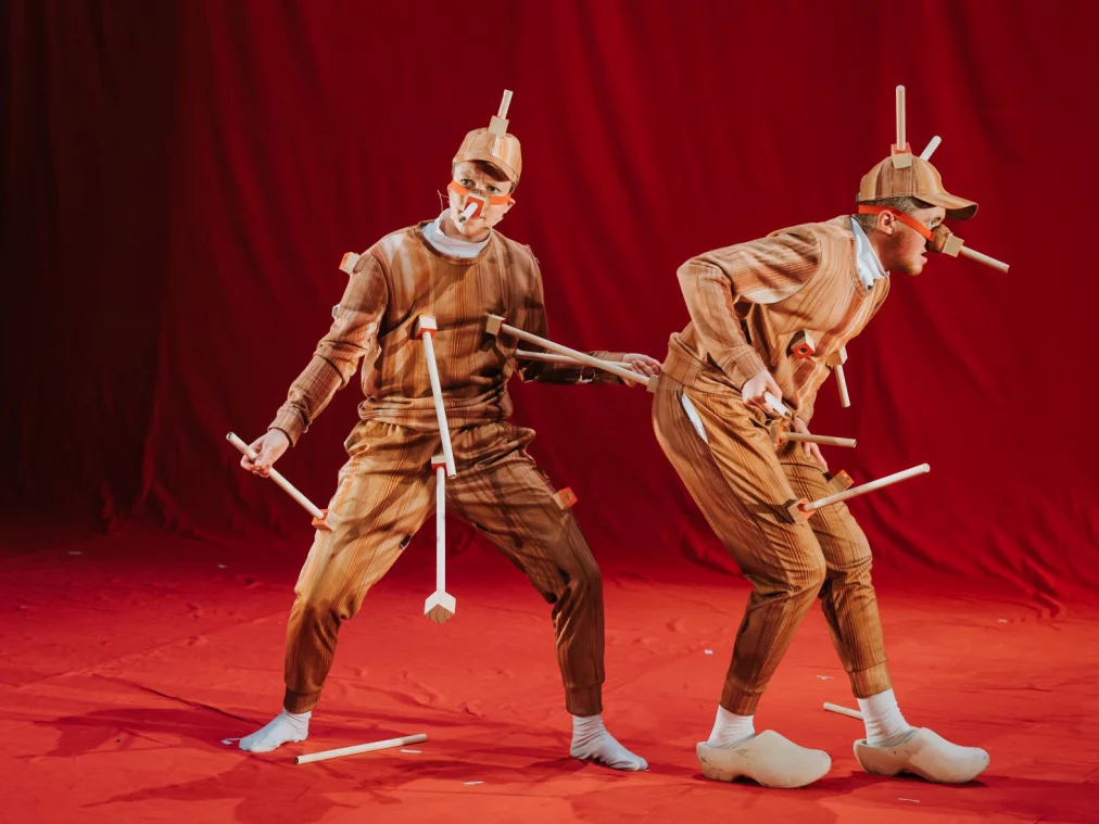 Rosana and Ivor stand on a stage draped in bright red fabric.   Rosana stands facing us, with a quizzical expression on their face. A wooden appendage protruding from Rosana’s nipple socket connects to Ivor’s arse socket.