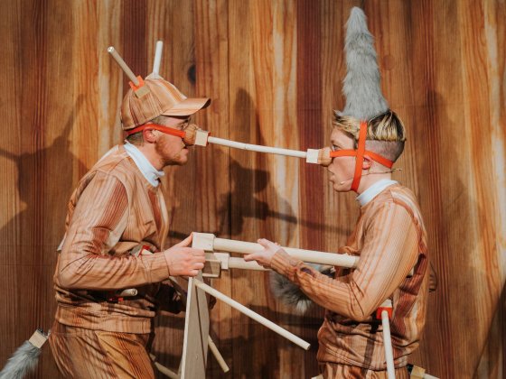 Two performers: Ivor MacAskill (he/him) and Rosana Cade (they/them), are shown in profile from the waist up, facing each other. They wear fake wooden tracksuits and caps with real wooden appendages protruding from them.